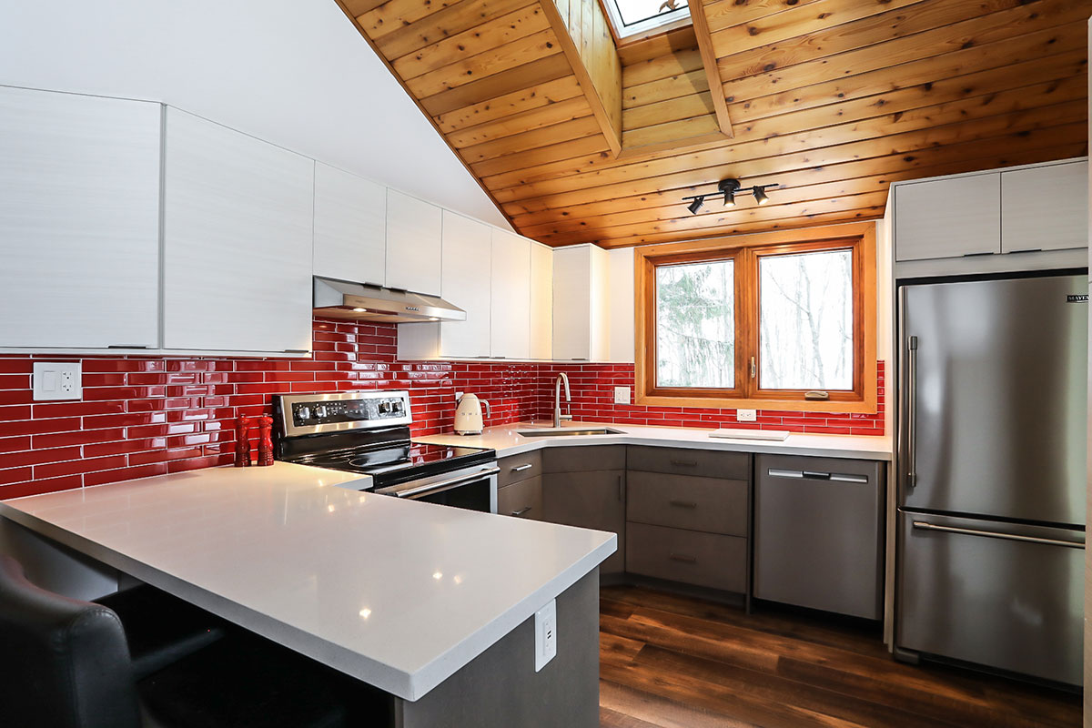 Kitchen renovation featuring two-toned white and warm grey cabinets, clean white countertops, stainless steel appliances, natural wood flooring, and red tile backsplash for a pop of colour.