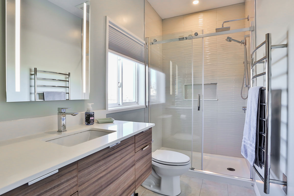 White bathroom design with tile flooring, large stand up shower with rain shower head, stainless steel accents including warming towel rack, and gleaming white vanity with sunken sink and teak cabinets
